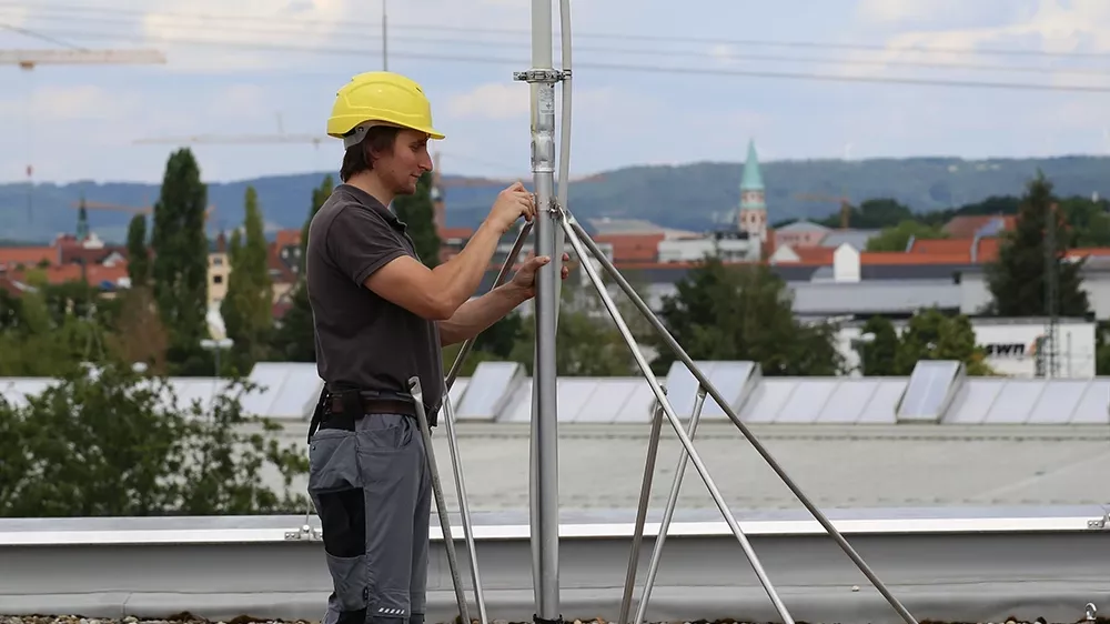 Person wearing a yellow helmet adjusts a metal mast with support rods on a gravel rooftop, buildings visible in the background.