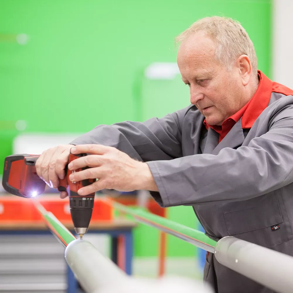 Worker in a grey lab coat using a cordless drill to fasten a metal rod in a workshop.