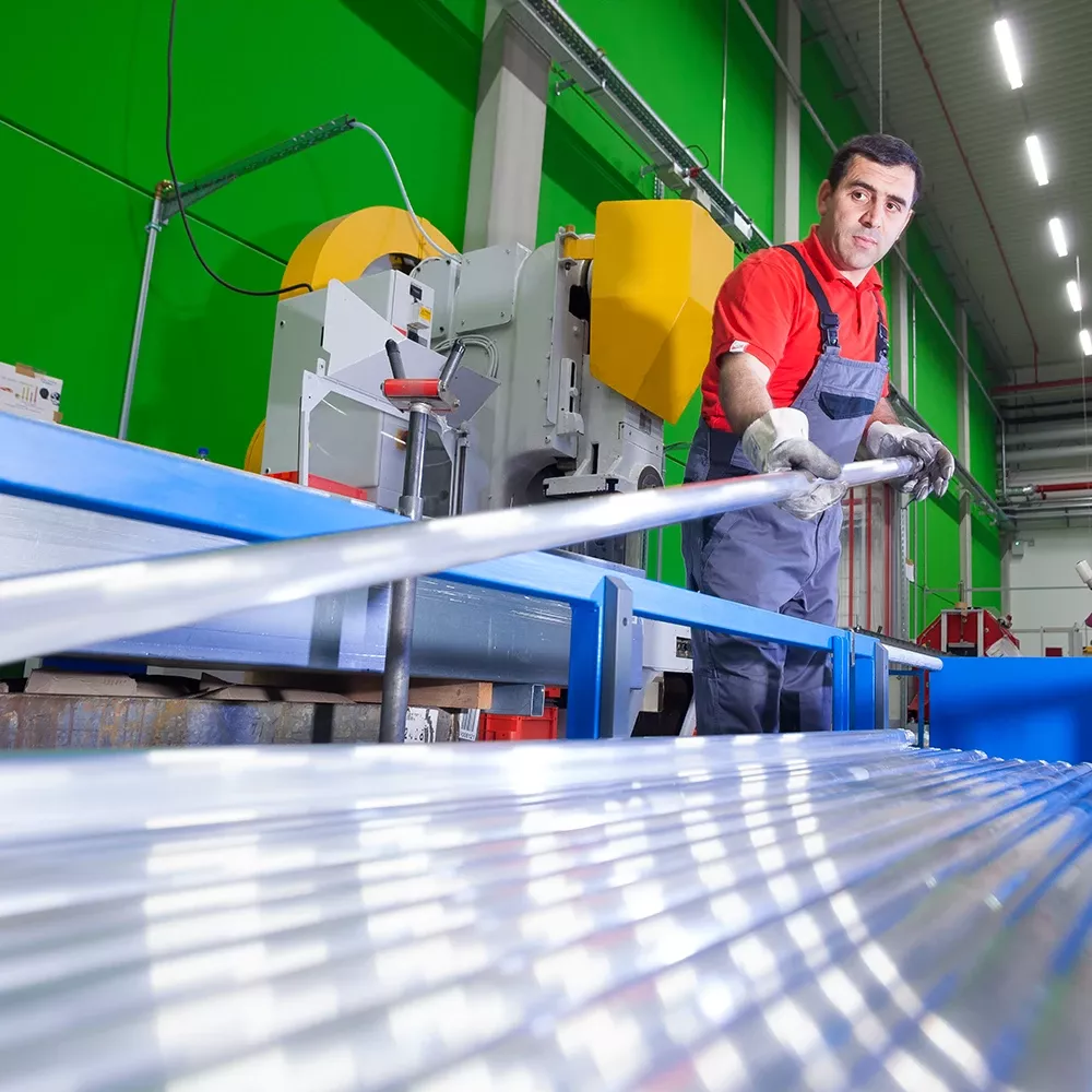 Worker in red shirt and overalls guiding a metal rod through a cutting machine in a factory.