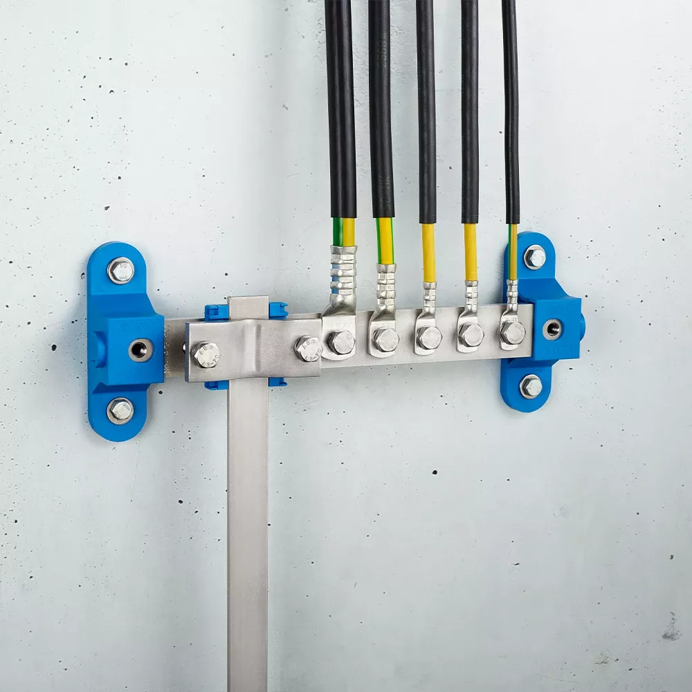 Wall-mounted grounding bar with metal clamps, connected black and yellow-green cables, and blue mounting brackets on a light gray wall.
