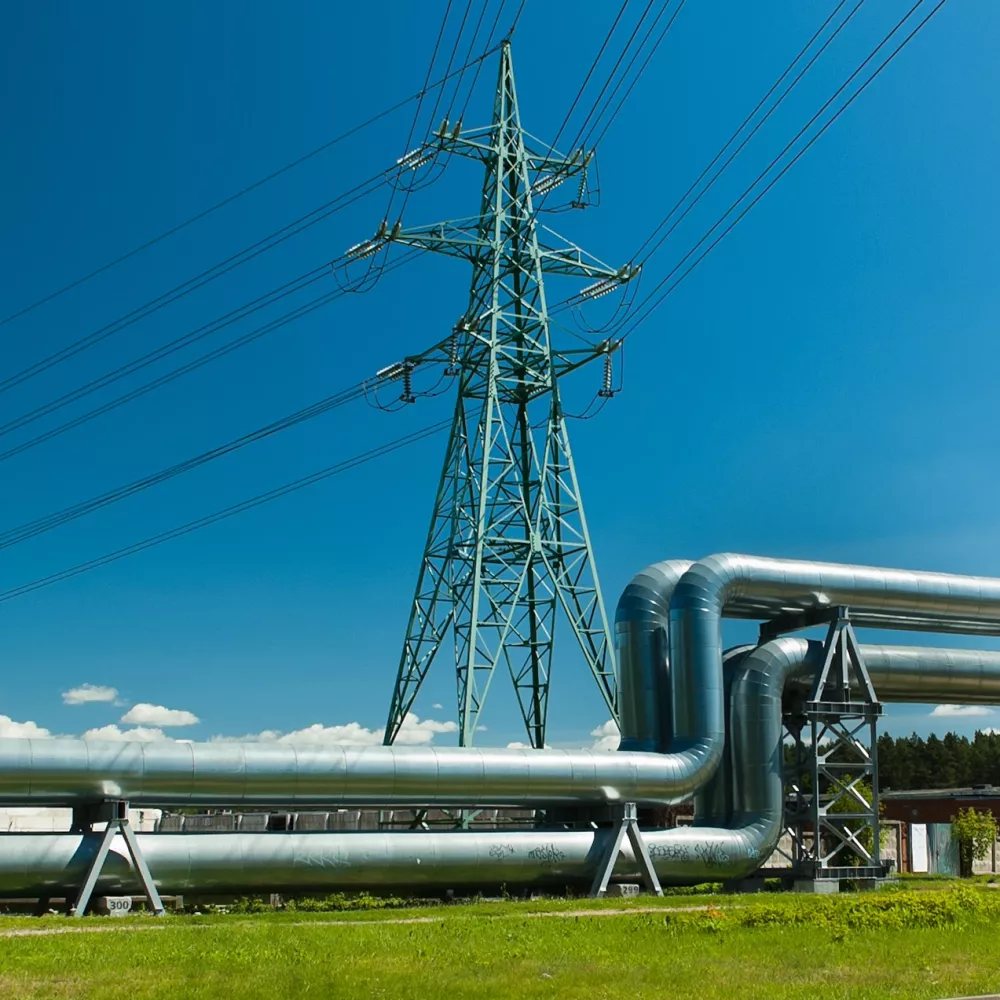 Industrial pipeline installation beneath a high-voltage transmission tower with overhead power lines under a clear blue sky.