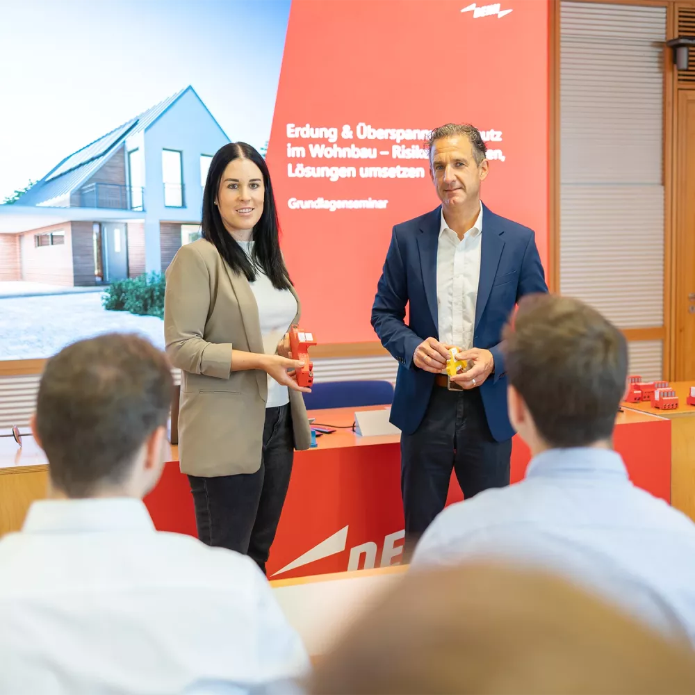 Two presenters holding devices stand in front of a screen reading “Erdung & Überspannungsschutz im Wohnbau – Grundlagenseminar” while speaking to an audience.
