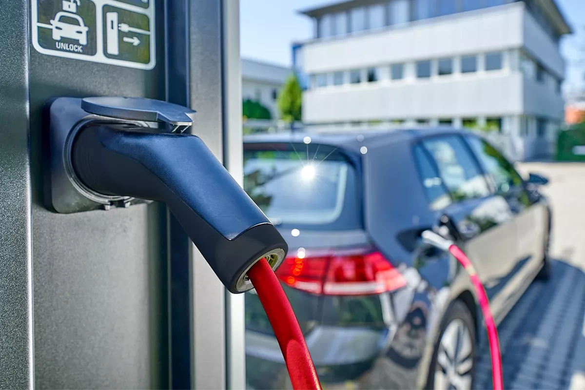 Close-up of an electric vehicle charging connector with red cable plugged into a charging station, car in background.